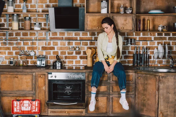 Young repairwoman sitting on table at kitchen and looking at oven — Stock Photo