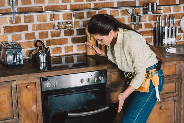 Confused young repairwoman looking at broken oven with smoke inside — Stock Photo