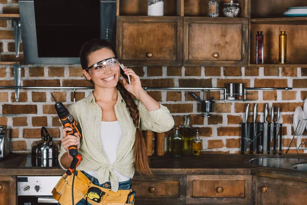 Счастливая молодая ремонтница в защитных очках с силовой дрелью разговаривает по телефону — стоковое фото