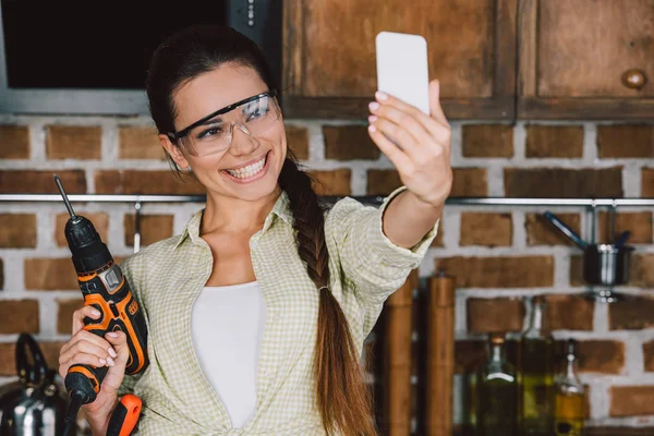 Happy young repairwoman in protective glasses with power drill taking selfie — Stock Photo