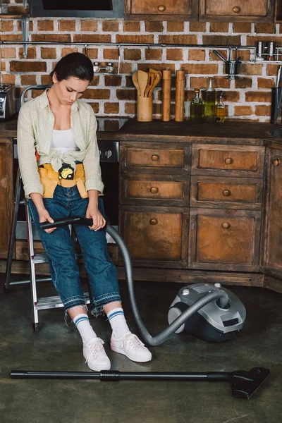 Sad young repairwoman looking at broken vacuum cleaner while sitting on stepladder at kitchen — Stock Photo