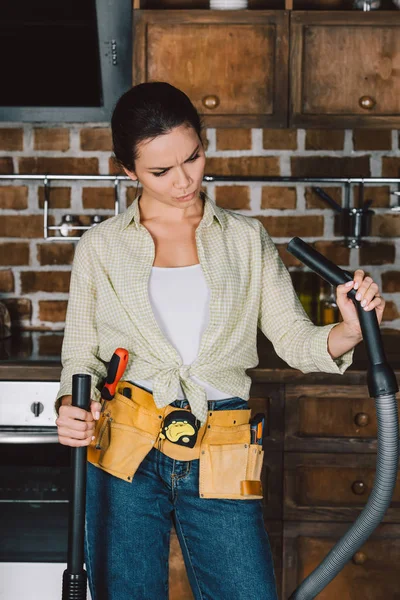 Confused young repairwoman looking at vacuum cleaner at kitchen — Stock Photo