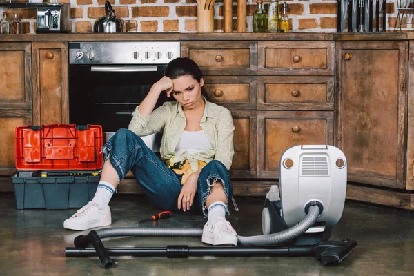 Depressed young woman sitting on floor of kitchen with vacuum cleaner — Stock Photo