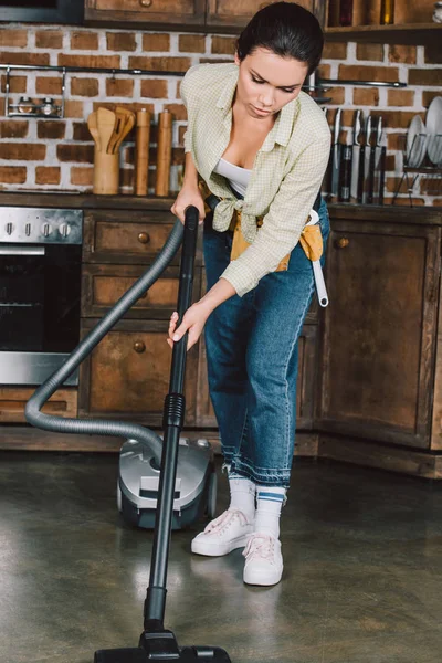 Mujer joven seria con cinturón de herramientas utilizando aspiradora en la cocina - foto de stock