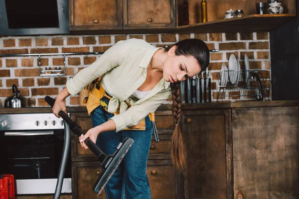 Confus jeune femme avec ceinture d'outils regardant tuyau d'aspirateur à la cuisine — Photo de stock