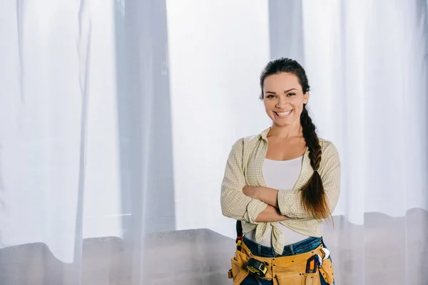 Hermosa joven reparadora con cinturón de herramientas y brazos cruzados mirando a la cámara - foto de stock