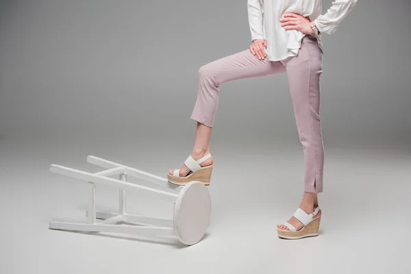 Cropped image of woman posing with leg on flipped over chair on grey background — Stock Photo