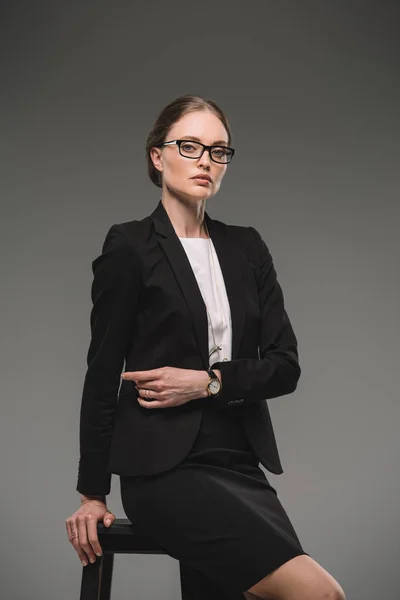 Confident businesswoman in eyeglasses looking at camera and sitting on chair isolated on grey background — Stock Photo