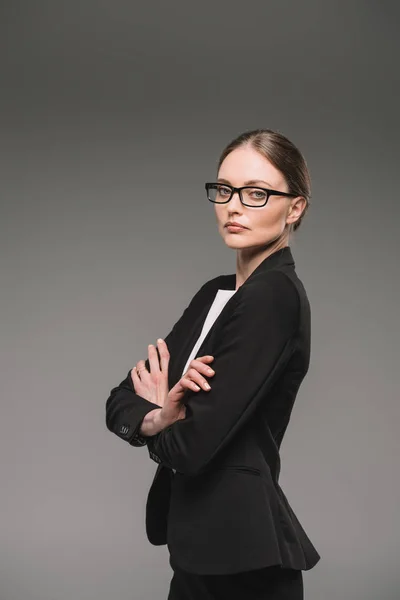 Empresaria confiada en gafas de pie con brazos cruzados aislados sobre fondo gris - foto de stock