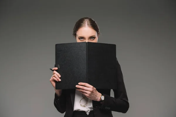 Серьезная деловая женщина, прикрывающая лицо учебником и смотрящая на камеру, изолированную на сером фоне — стоковое фото