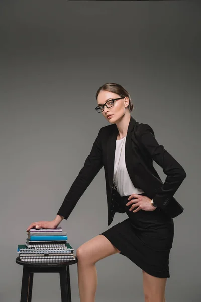 Female teacher in eyeglasses standing near stack of textbooks on chair on grey background — Stock Photo