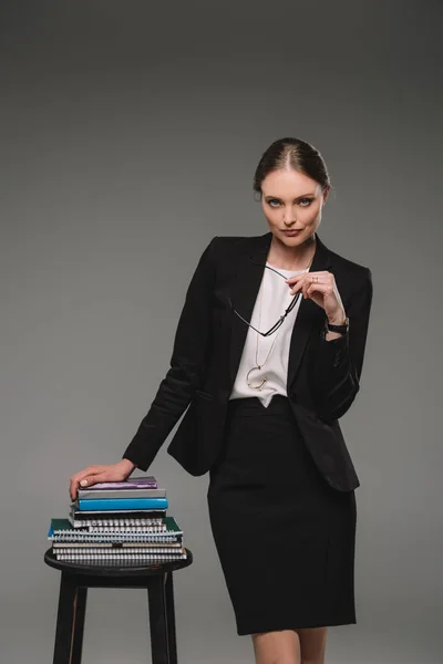 Confident female teacher standing near chair with stack of textbooks on grey background — Stock Photo