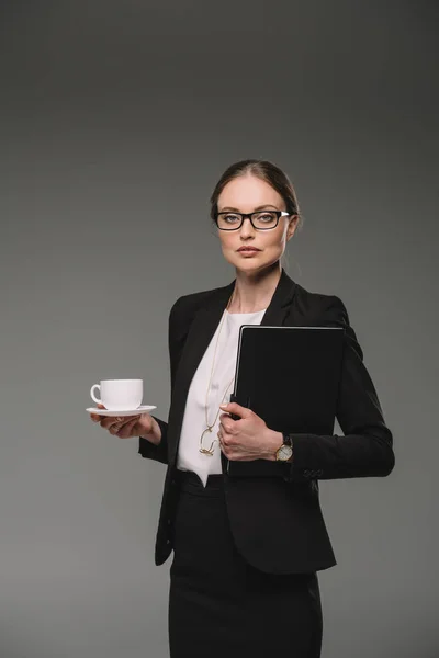 Серйозна бізнес-леді в окулярах тримає чашку кави і підручник ізольовані на сірому фоні — стокове фото