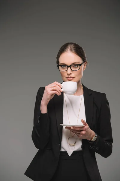 Atractiva mujer de negocios en anteojos bebiendo café de taza aislada sobre fondo gris - foto de stock