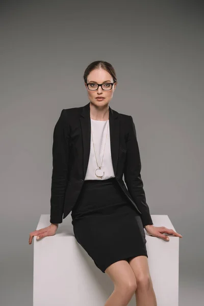 Séduisante femme d'affaires en lunettes assise sur cube sur fond gris — Photo de stock