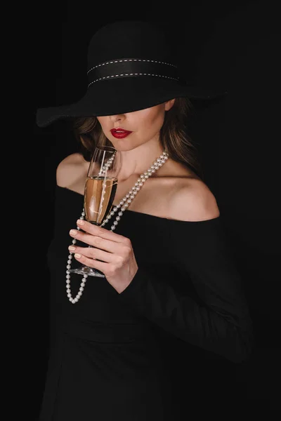 Mujer elegante con los ojos cubiertos por paja negro celebración copa de champán aislado sobre fondo negro - foto de stock