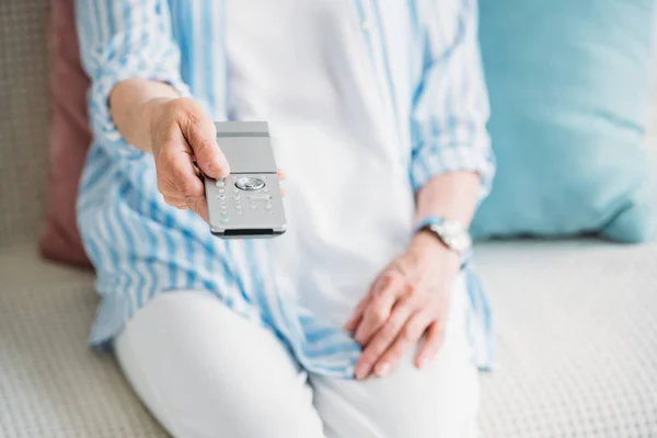 Partial view of senior woman with remote control watching tv on sofa at home — Stock Photo