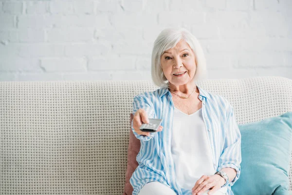 Portrait of smiling senior woman with remote control watching tv on sofa at home — Stock Photo