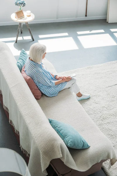 Visão de alto ângulo da mulher idosa com controle remoto assistindo tv no sofá em casa — Fotografia de Stock