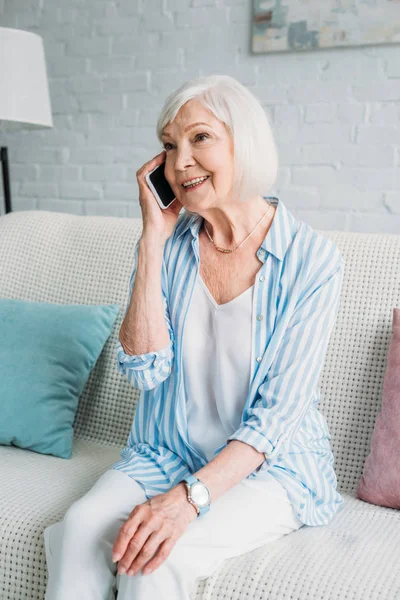 Портрет улыбающейся пожилой женщины, сидящей на диване и разговаривающей на смартфоне дома — стоковое фото