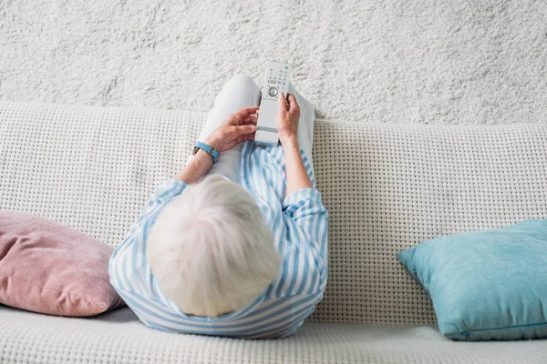 Вид сверху пожилой женщины с пультом, смотрящей телевизор на диване дома — стоковое фото