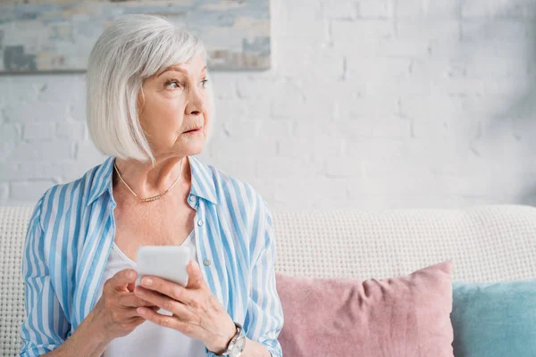 Portrait de femme âgée coûteuse avec smartphone regardant ailleurs reposant sur le canapé à la maison — Photo de stock