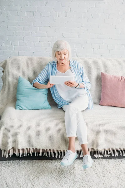 Mujer mayor que usa la tableta mientras descansa en el sofá en casa - foto de stock