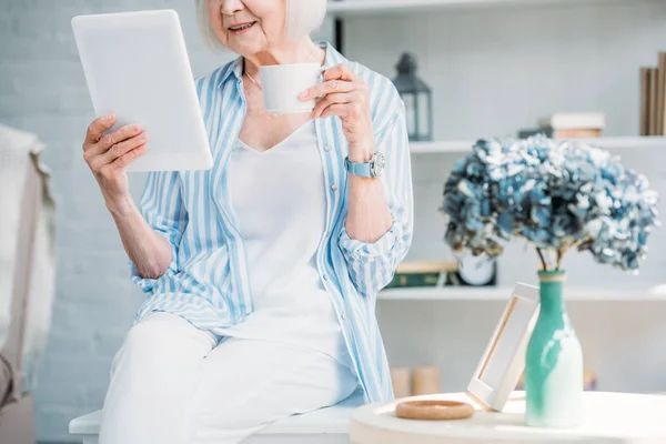 Vue partielle de la femme âgée avec tasse de café en utilisant une tablette à la maison — Photo de stock