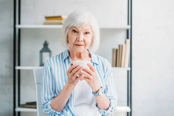 Porträt einer Seniorin mit einer Tasse aromatischen Kaffees in der Hand, die zu Hause in die Kamera blickt — Stockfoto