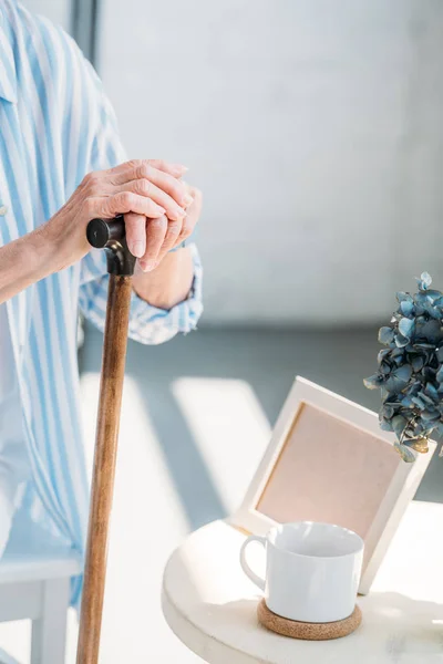 Vista parziale della donna anziana con bastone da passeggio al tavolino con tazza di caffè e cornice fotografica vuota — Foto stock