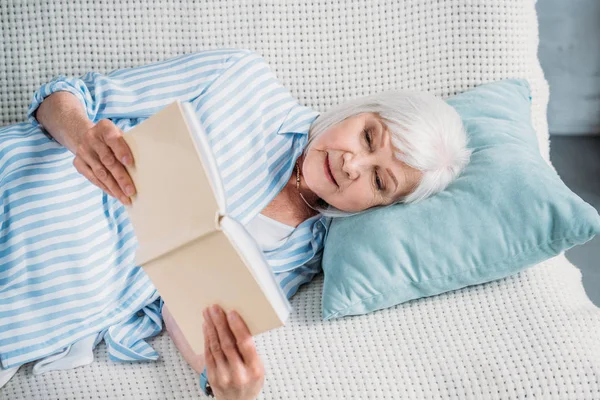 Retrato de la mujer mayor sonriente leyendo libro mientras descansa en el sofá en casa - foto de stock