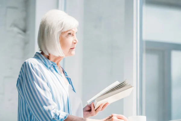 Vue latérale de la femme aux cheveux gris avec livre et tasse de café debout au rebord de la fenêtre à la maison — Photo de stock