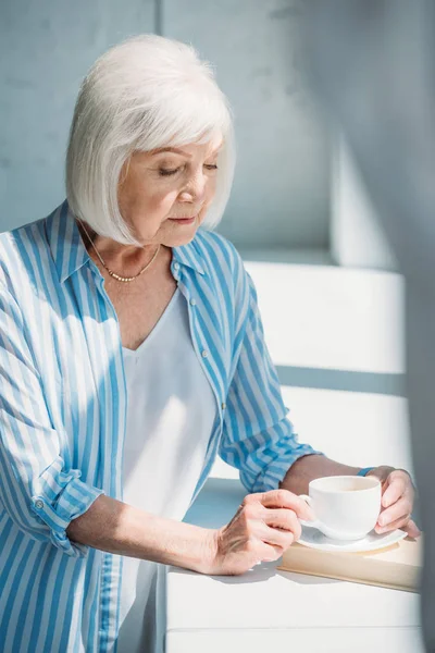 Pensive grey hair woman with cup of coffee standing at windowsill with book at home — Stock Photo
