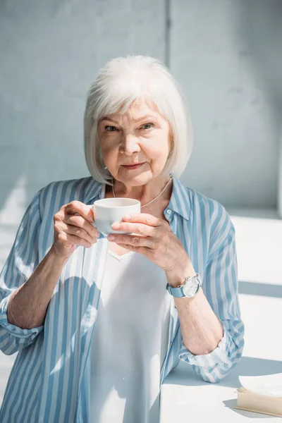 Porträt einer grauhaarigen Frau mit einer Tasse Kaffee, die zu Hause auf der Fensterbank steht — Stockfoto