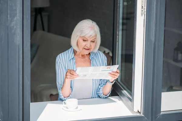 Senior woman standing at windowsill with cup of aromatic coffee and reading newspaper at home — Stock Photo