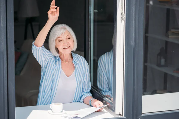 Пожилая женщина приветствует кого-то, стоя на подоконнике с газетой и чашкой ароматного кофе дома — стоковое фото