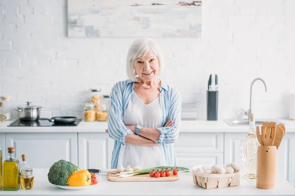 Ritratto di anziano sorridente con le braccia incrociate in piedi al bancone con verdure fresche in cucina — Foto stock