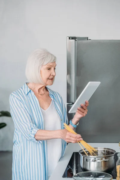 Side view of senior woman with digital tablet cooking macaroni in kitchen at home — Stock Photo