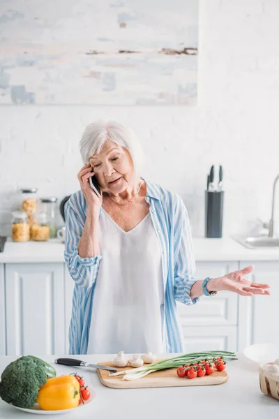 Retrato de la mujer mayor hablando en el teléfono inteligente, mientras que de pie en el mostrador con verduras frescas en la tabla de cortar en la cocina - foto de stock