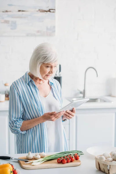 Donna anziana con tablet digitale in piedi al bancone con verdure fresche in cucina — Foto stock