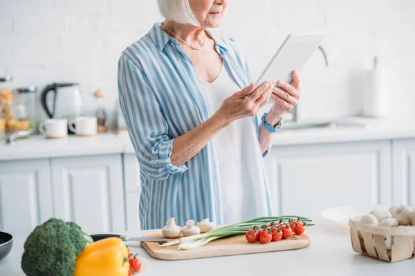 Частичный вид пожилой женщины с цифровым планшетом стоя у прилавка со свежими овощами на кухне — стоковое фото