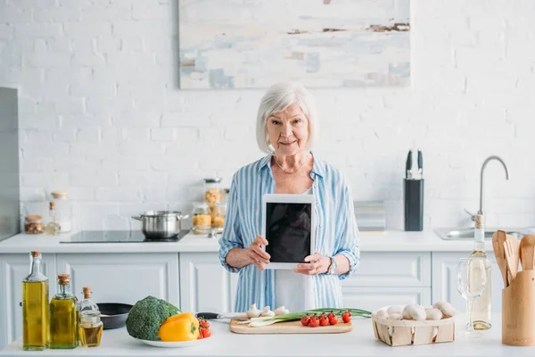 Портрет пожилой женщины, показывающей планшет с чистым экраном, стоя за прилавком со свежими овощами на кухне — стоковое фото