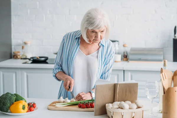 Retrato de la señora mayor que busca receta en el libro de cocina mientras cocina la cena en el mostrador en la cocina - foto de stock