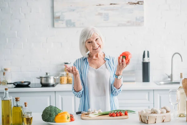 Портрет усміхненої сірої волосинки з болгарським перцем в руці, що стоїть за прилавком на кухні — стокове фото