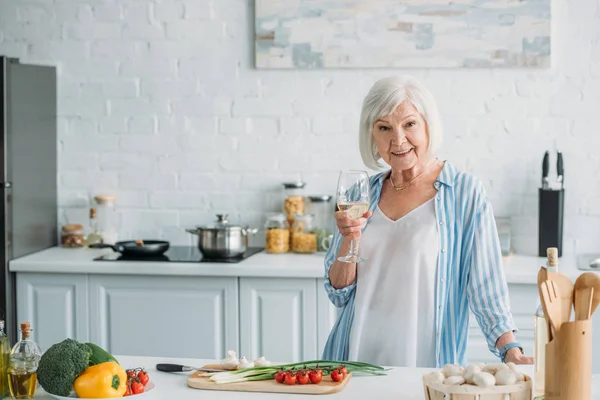 Портрет усміхненої старшої леді з келихом вина, що стоїть за лічильником зі свіжими овочами на кухні — стокове фото