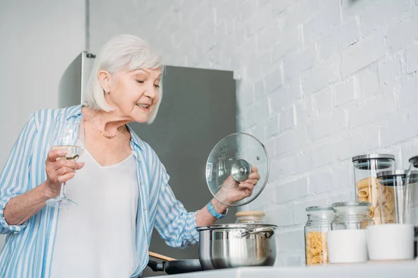 Vista laterale della signora anziana con bicchiere di vino pentola di controllo sul fornello in cucina — Foto stock