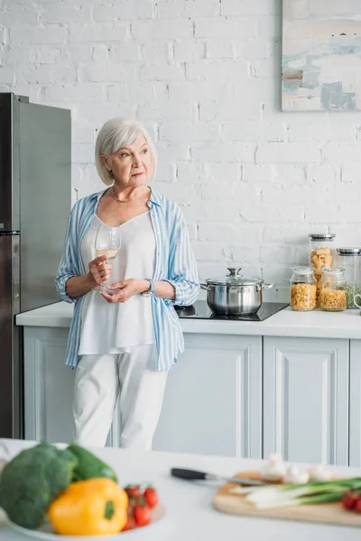 Selective focus of fresh vegetables on counter and senior woman with glass of wine standing at stove in kitchen — Stock Photo