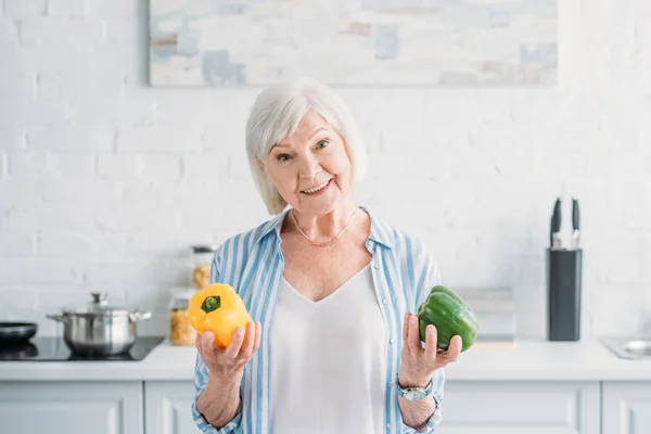 Retrato de la señora mayor sonriente con pimientos frescos en las manos en la cocina - foto de stock