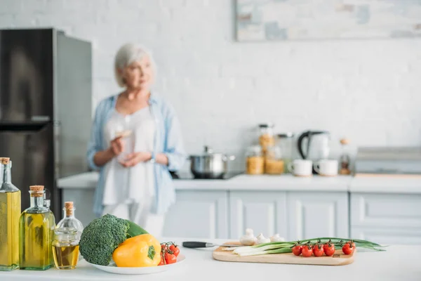 Enfoque selectivo de verduras frescas en el mostrador y la mujer mayor con vaso de vino de pie en la estufa en la cocina — Stock Photo