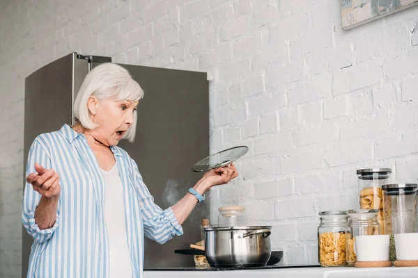 Vista lateral de la señora mayor sorprendida chequeando cacerola en estufa en la cocina — Stock Photo